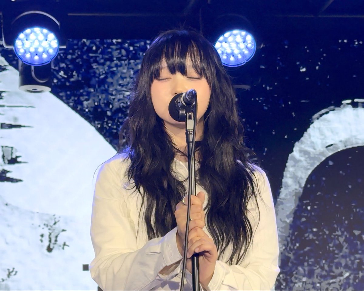A young woman with long dark wavy hair and bangs stands at a microphone with her eyes closed, appearing to sing. She wears a white blouse, and bright blue stage lights illuminate the dark background.