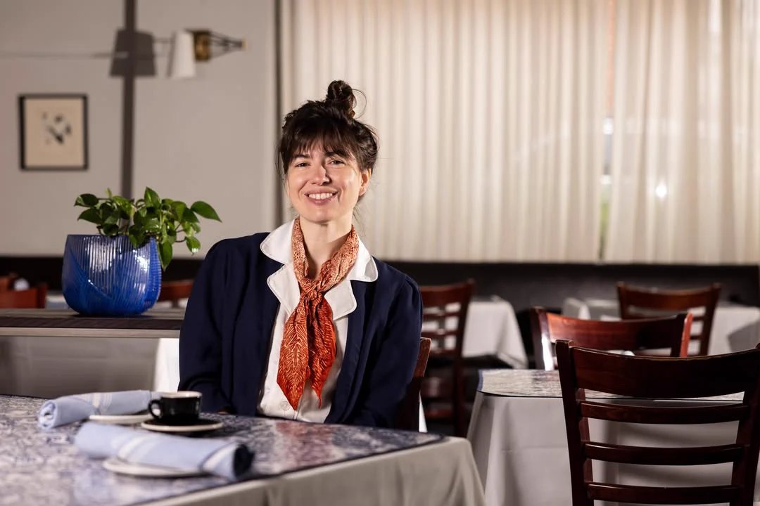 A person with a bun hairstyle is seated at a table in a restaurant, smiling. They are wearing a blue jacket and an orange scarf. A blue pot with a green plant is on the table beside a cup and saucer. The room has a calm ambiance with light-colored curtains in the background.