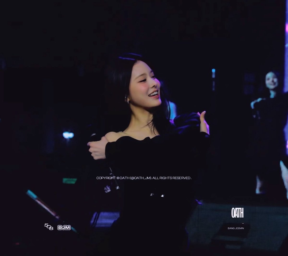 A woman with dark hair smiles on stage at a concert, wearing a black off-shoulder top, with a watermark reading 'COPYRIGHT © OATH (@OATH_JM). ALL RIGHTS RESERVED.'