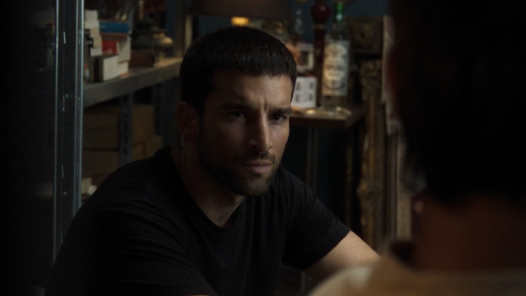 A scene featuring a man with a beard and short dark hair, wearing a black t-shirt, sitting and looking intently at someone off-camera.