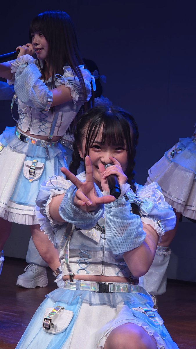 A young female performer in a light blue and white idol costume smiles and holds up a peace sign toward the camera while holding a microphone on stage.