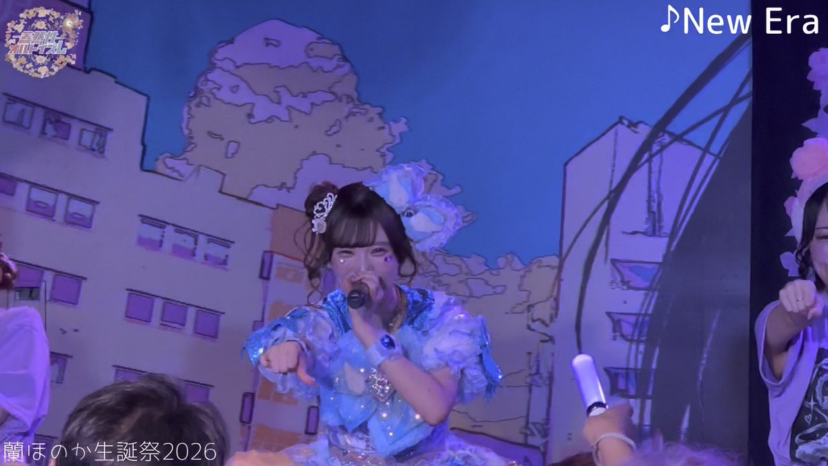 A performer with brown hair in a high ponytail sings into a microphone on stage, wearing a frilly blue dress; the background features an anime-style illustration, with text reading '蘭 ほのか生誕祭2026' in the bottom left and '♪New Era' in the top right.