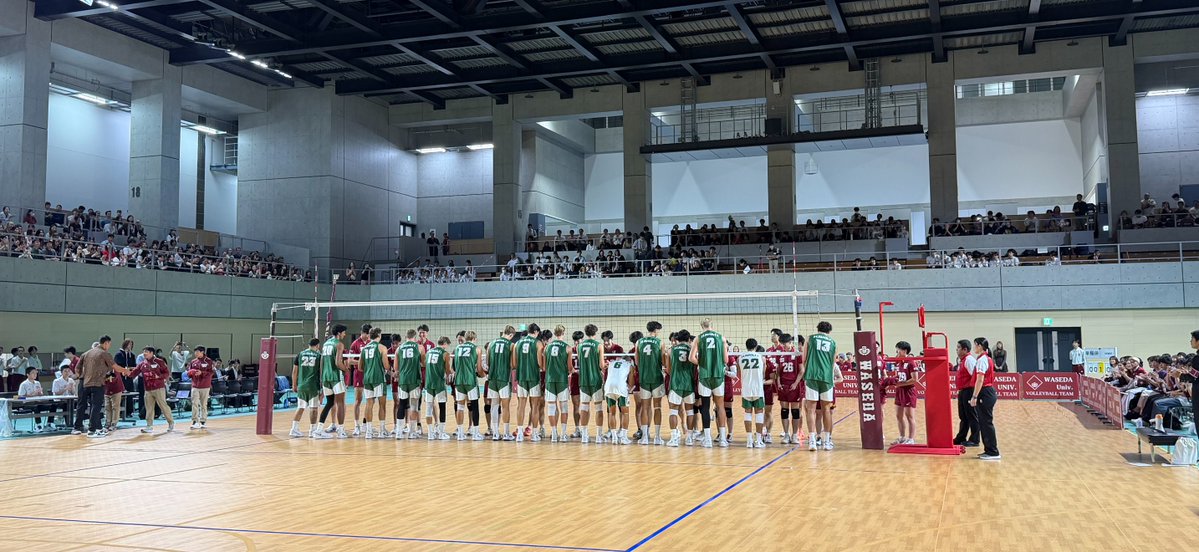 Two men's volleyball teams, one in green and one in red, line up at the net to shake hands in a large gymnasium, with signage reading 'WASEDA' and 'WASEDA MEN'S VOLLEYBALL TEAM' visible.