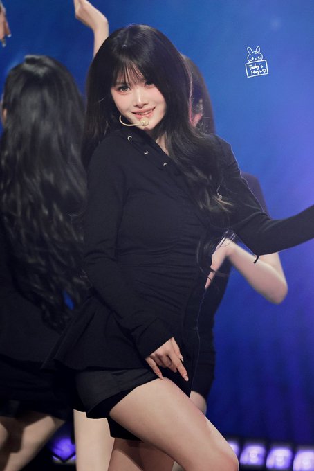K-pop performance photo of a young woman with long dark hair in a black long-sleeved top and skirt, wearing a headset microphone, performing on stage with backup dancers.