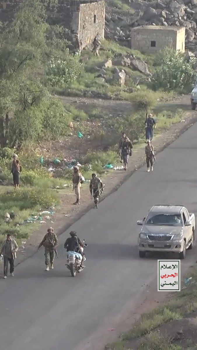 An aerial view of armed soldiers walking along a road and rocky hillside near buildings, with a white pickup truck and motorcycle, labeled in Arabic as Yemeni media.