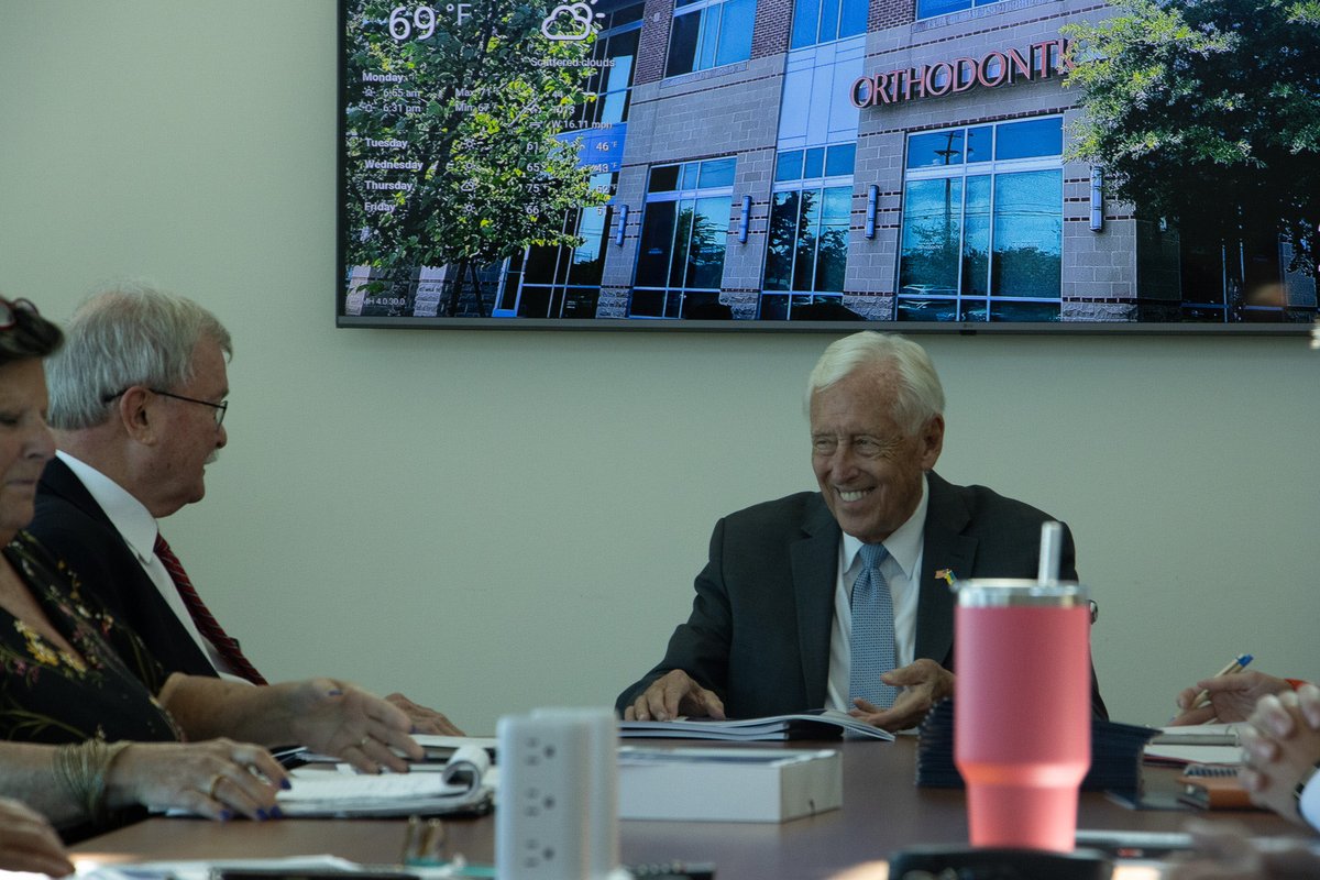 Several older adults in business attire sit around a conference table during a meeting. A smiling man sits at the center, with papers and writing materials on the table. A wall-mounted screen behind them shows a building and weather information, and a pink tumbler is in the foreground. Text appears on the screen.
