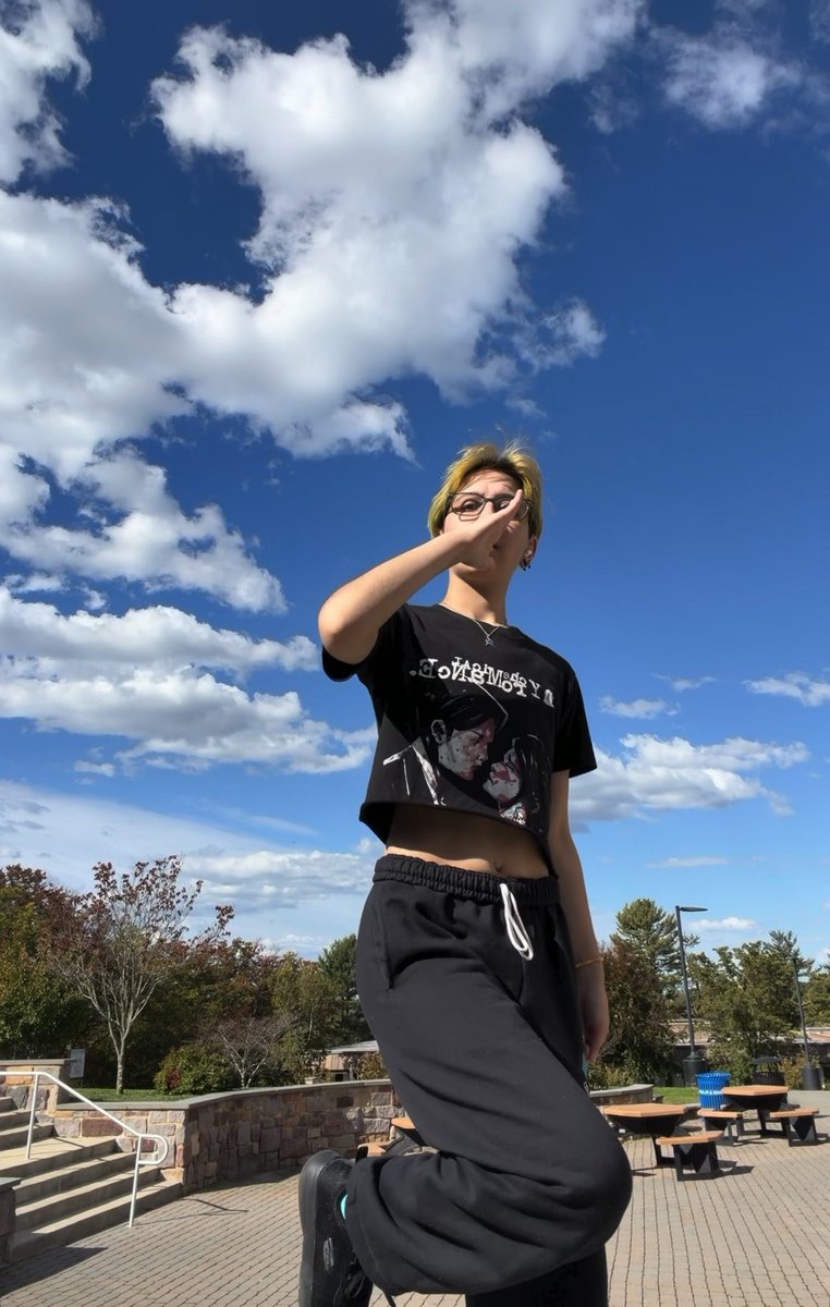 A person with blonde hair and glasses wearing a black graphic t-shirt and dark sweatpants stands outdoors on a sunny day with a blue sky and scattered clouds, posing with a hand gesture near their face.