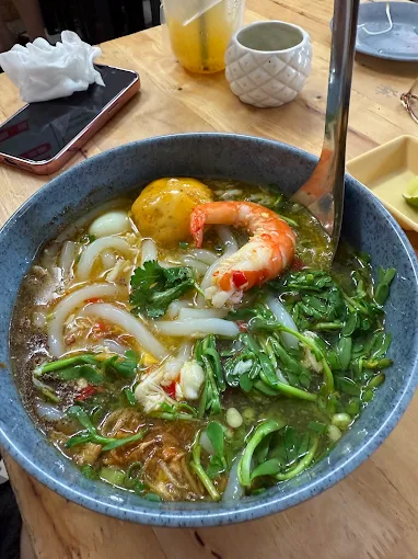 A steaming bowl of Vietnamese crab noodle soup (bánh canh cua) topped with a whole shrimp, fresh herbs, and thick rice noodles sits on a wooden table beside a phone and a pineapple-shaped cup. The caption highlights the satisfying, hearty nature of this comforting breakfast dish.