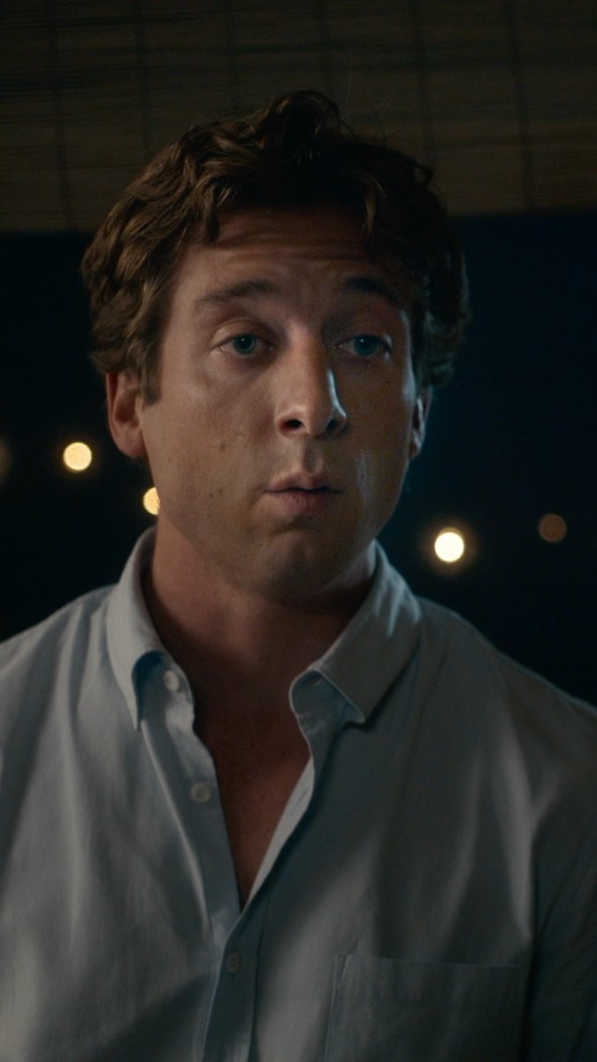 A young man with tousled brown hair and blue eyes, wearing an unbuttoned light blue shirt, stares directly at the camera with a slightly pensive expression against a dark background with blurred warm lights. The image appears to be promotional material for "The Social Reckoning," the companion piece to The Social Network, suggesting the man pictured is an actor portraying a key figure in the film.