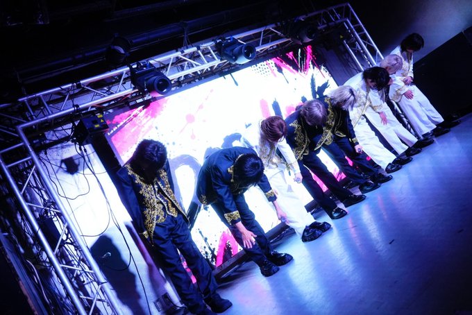Members of a performance group bow on stage under dramatic blue and pink lighting in elaborate black and white costumes with gold accents.