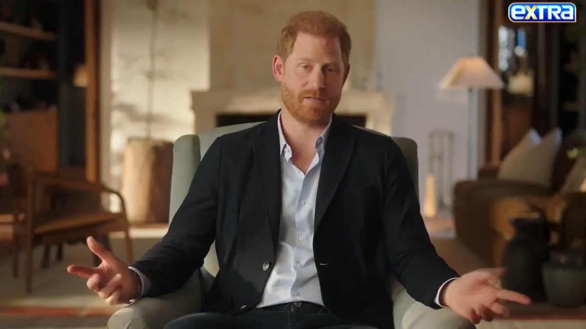 Prince Harry sits in a light-colored armchair during an interview, gesturing with both hands open. The 'extra' TV show logo appears in the top right corner.