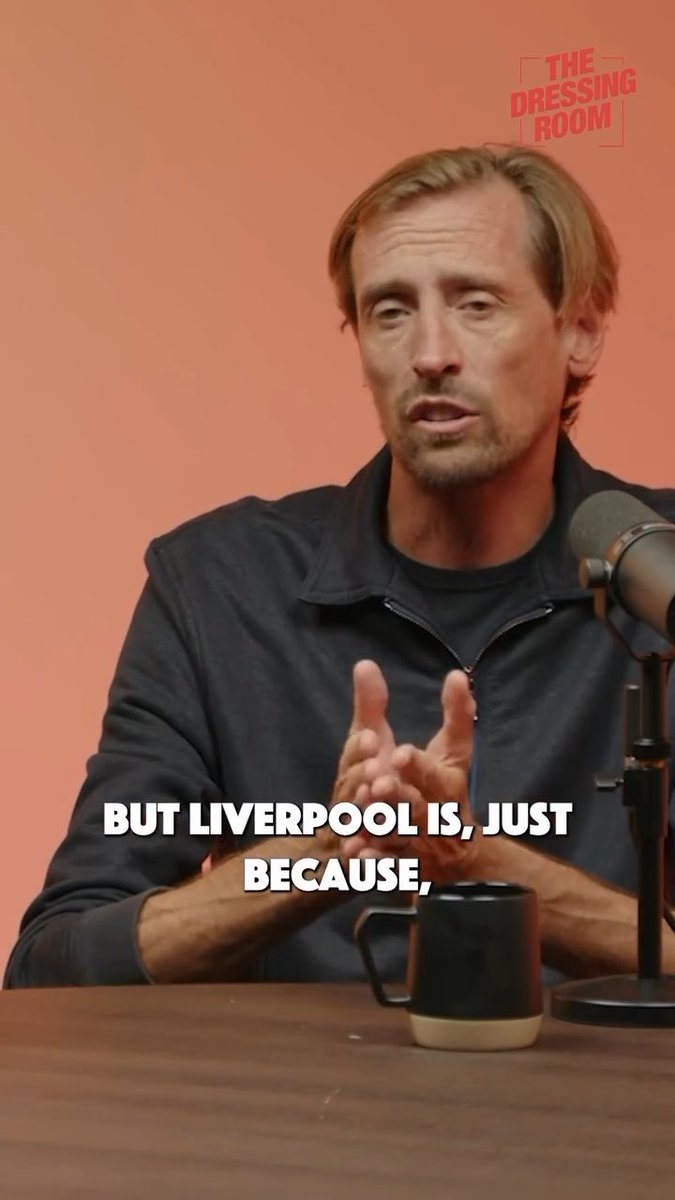 Peter Crouch speaks into a microphone at a table with a black mug, against a peach-colored background. A red “THE DRESSING ROOM” logo appears in the top-right corner, and the bold white caption reads, “BUT LIVERPOOL IS, JUST BECAUSE,” matching his reflections on his time at Liverpool and what the city means to him.