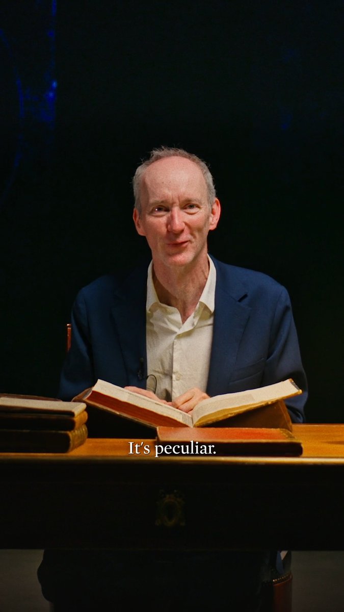 A man with light hair, wearing a blue blazer over a light shirt, sits at a wooden desk reading from an open book with stacked books beside him, against a dark background, with the subtitle 'It's peculiar.'