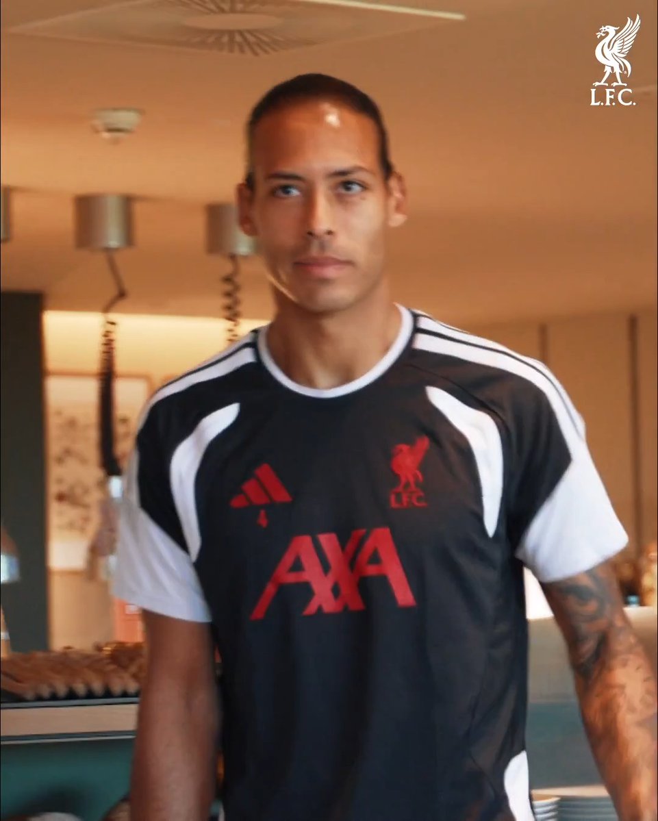 A behind-the-scenes shot shows a Liverpool player in a black Adidas training shirt with white shoulder stripes, the red Liverpool crest, and the red sponsor text “AXA” across the chest. The white Liverpool crest and “L.F.C.” appear in the top-right corner, matching the post reference “Redz Behind the Scenes” and the link “lfc.tv/4yeY4XF.”