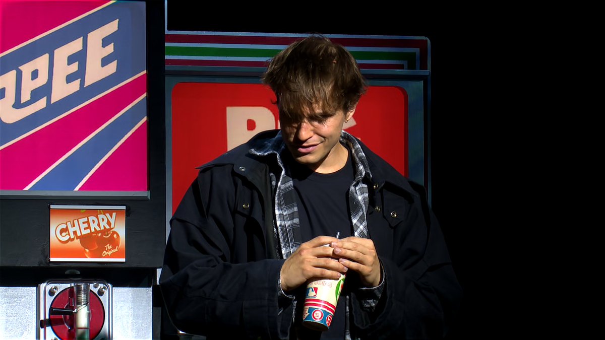 A young man with messy brown hair standing next to a vintage-style 'PEE' machine prop, holding a slushie cup, with a 'CHERRY' sign visible on the machine.