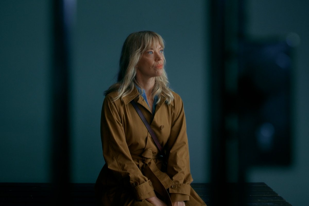 A blonde woman in a tan trench coat sits alone in a dimly lit, bluish room, her gaze cast upward with a sorrowful expression—evoking the loneliness mentioned in the post about a mother whose daughter is imprisoned. The melancholic atmosphere and solitary framing reinforce the emotional weight of "Where's Wanda?" Season 2, premiering October 21.