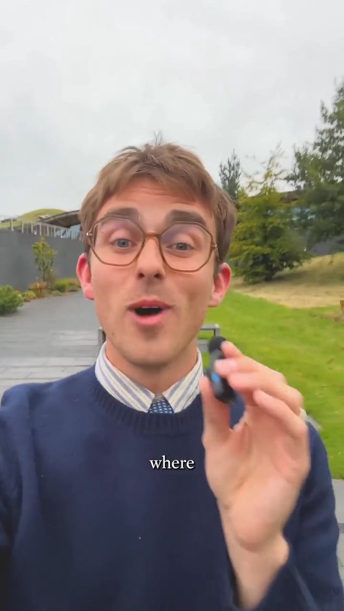 A young man with glasses and a blue sweater speaking outdoors, with the word 'where' displayed as a subtitle on the video.