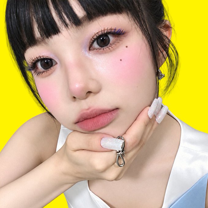 A close-up portrait of a young woman with short dark hair, bold colorful makeup, and long sparkly white nails resting her chin on her hand against a bright yellow background.