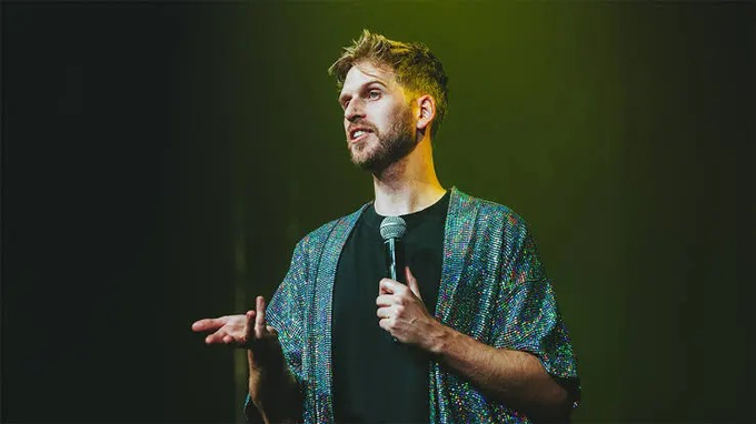 A young man with tousled brown hair and a short beard stands on a dimly lit stage holding a microphone, mid-gesture while performing stand-up comedy. He wears a black t-shirt under an iridescent, multicolored sequined kimono-style jacket that shimmers in the green stage lighting. The image accompanies a post jokingly accusing the comedian—appearing to be a foreign performer on stage—of being a "spy from Macron's leftist government," humorously playing on his international, possibly French, presence in Brazil.