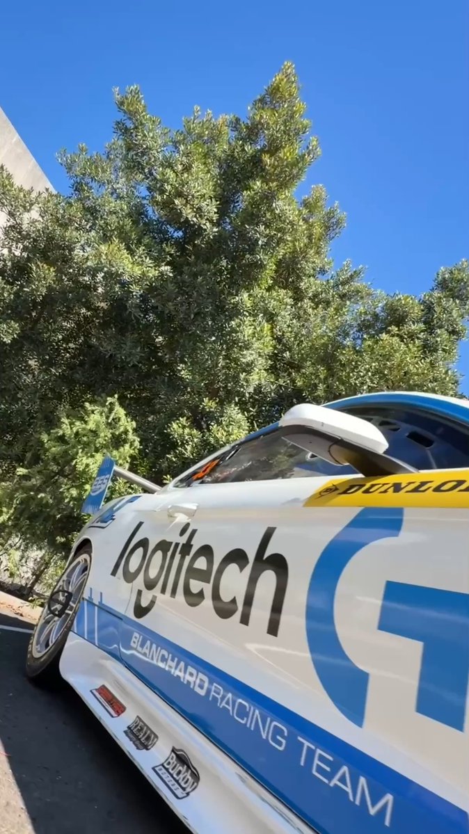 A white and blue racing car with 'logitech' and 'BLANCHARD RACING TEAM' decals, parked outdoors next to green trees.