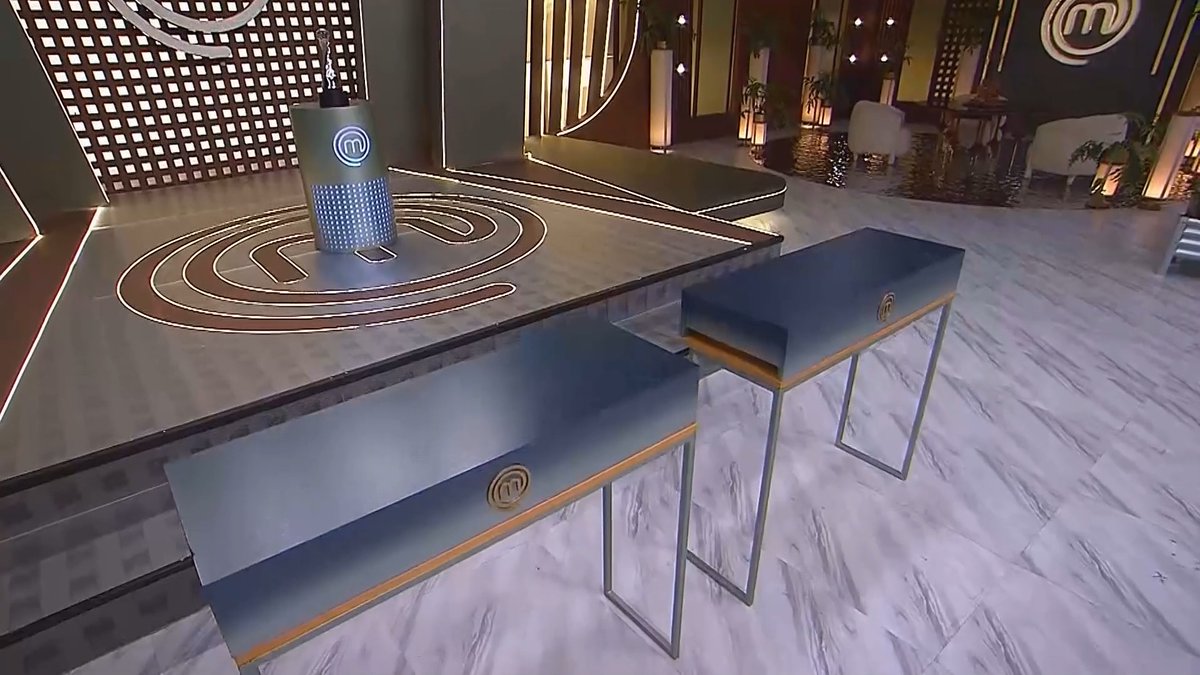 an elegantly lit studio set with marble floors, blue benches with MasterChef logos, and a cooking station marked with the MasterChef "M" emblem