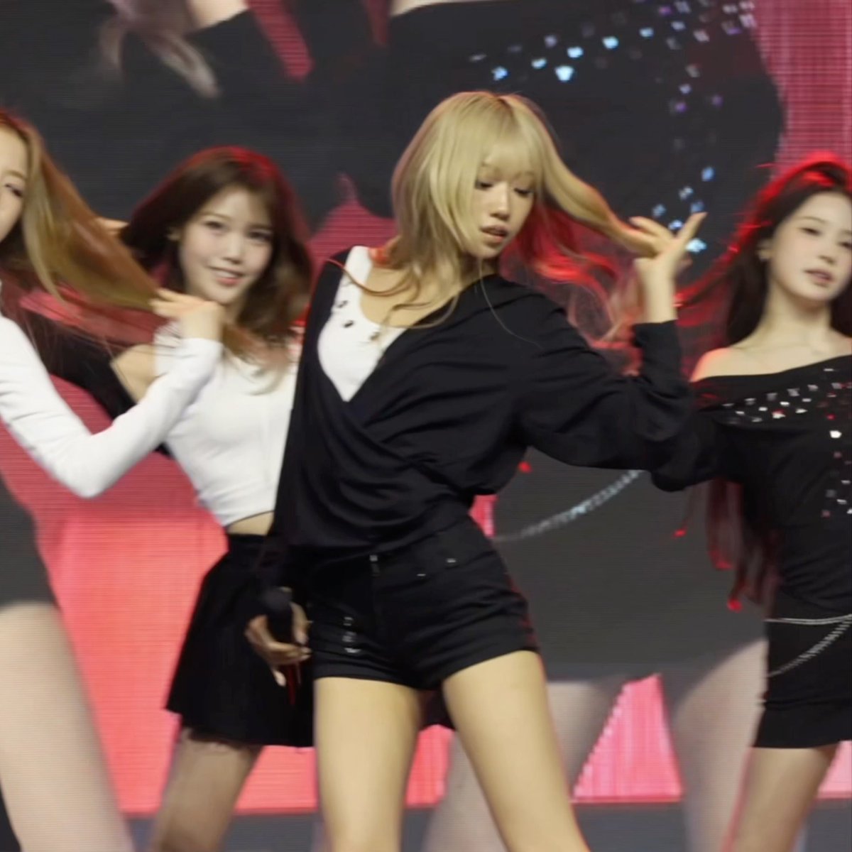 A group of female K-pop performers on stage in black outfits performing a choreographed dance routine under pink stage lighting.