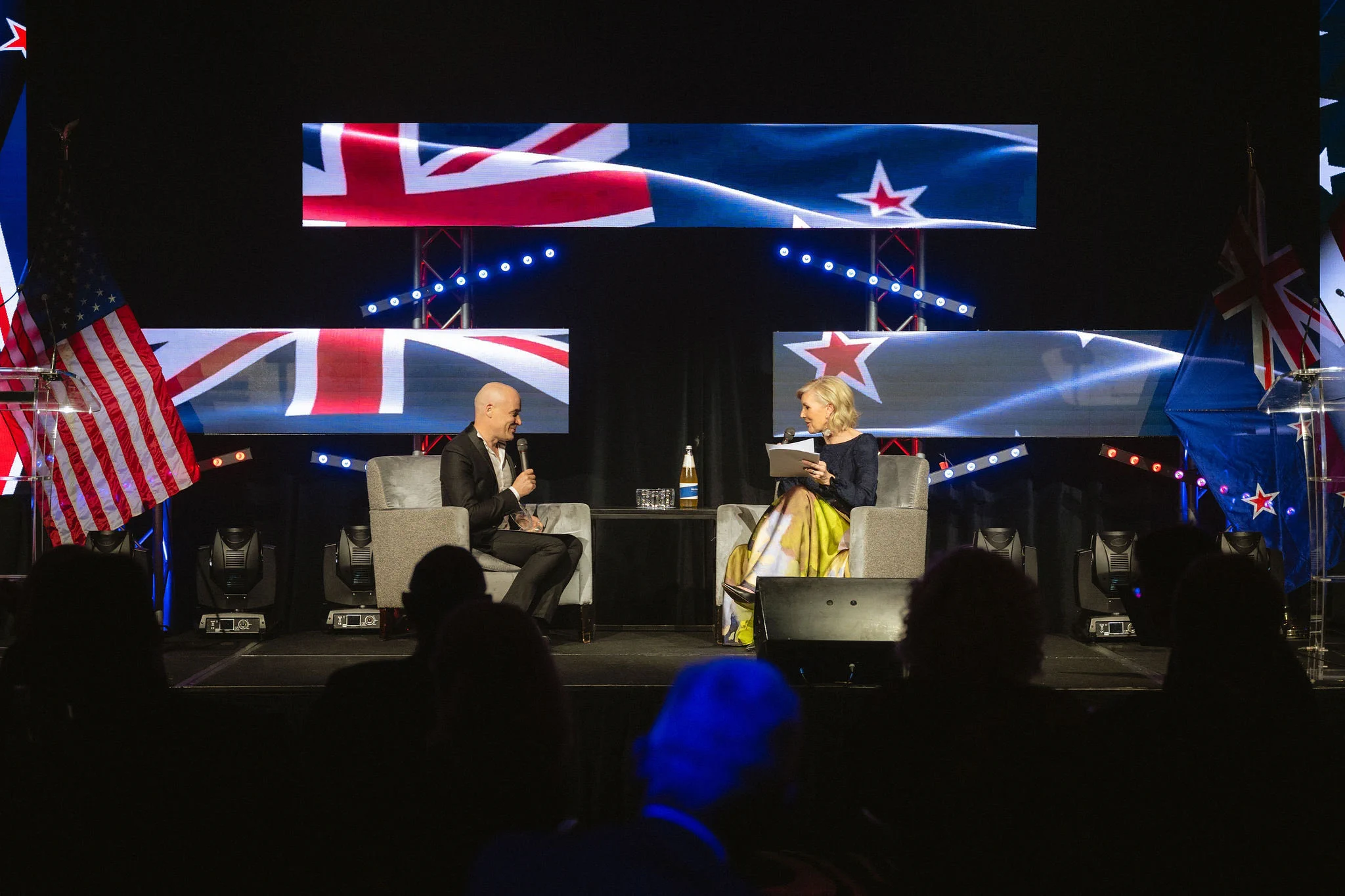 Professor Greg O’Grady and an interviewer sit in armchairs holding microphones, with U.S. and New Zealand flags displayed around the stage.