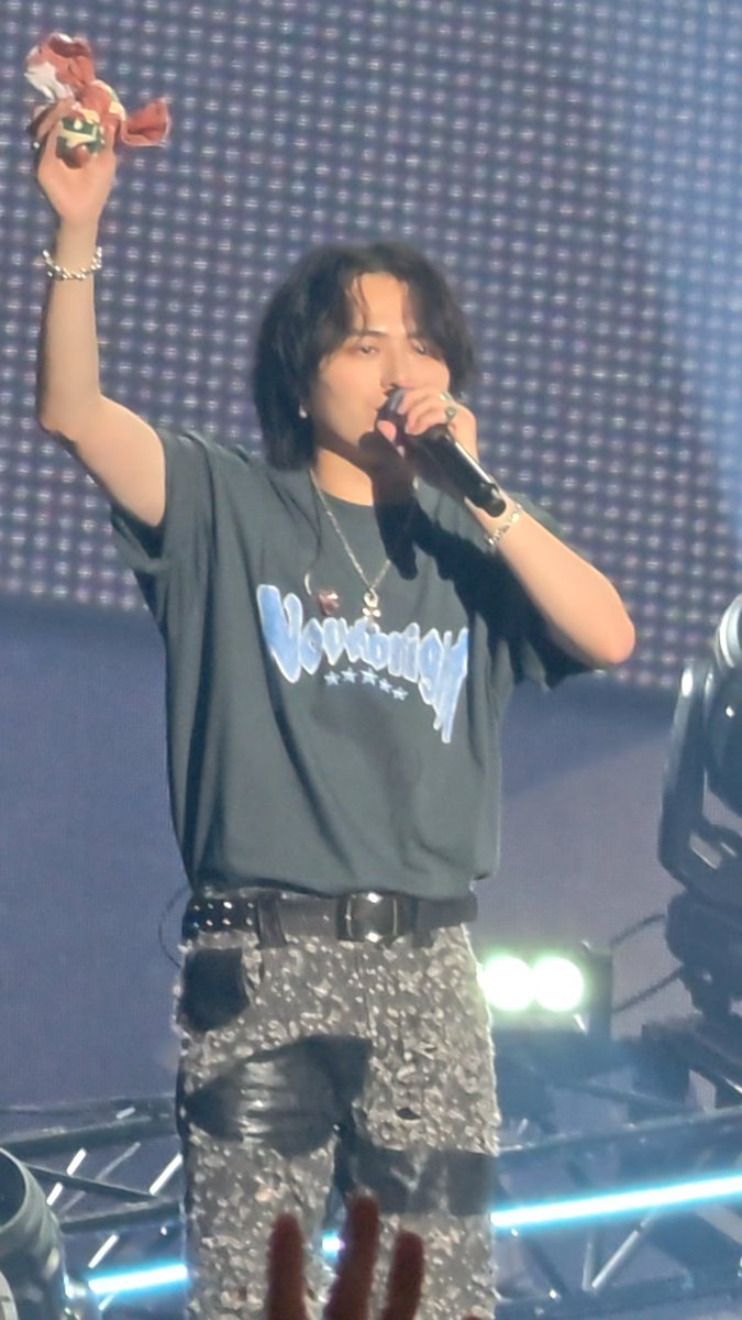 A concert photo of a performer with shoulder-length dark hair holding a microphone and a small bouquet, wearing a gray graphic t-shirt and floral-patterned pants on stage.