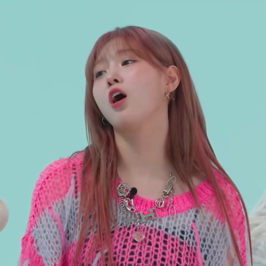 A young woman with long pinkish hair and a pink mesh top speaks against a light teal background.