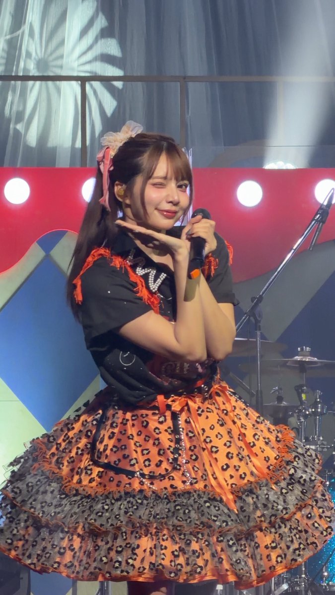 A female performer in an orange and black frilled dress with bow hair accessories holds a microphone on stage, winking at the camera.