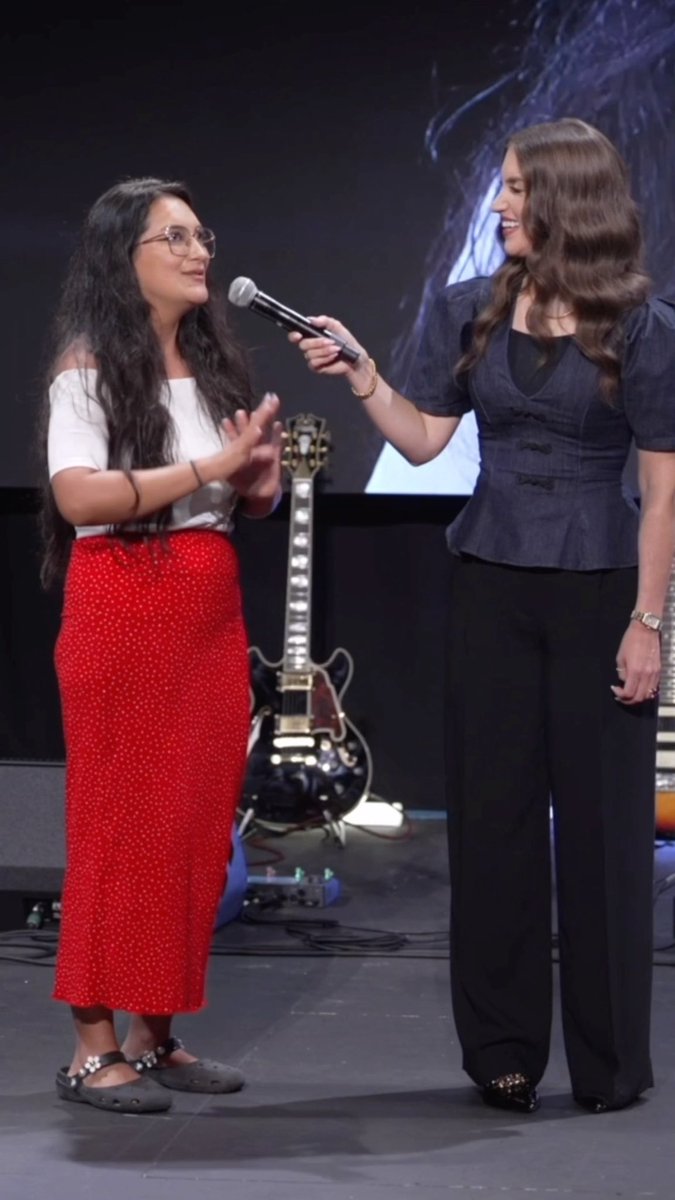 Two women stand on a stage with a guitar in the background; one with long dark hair wears a white top and red polka-dot skirt, while the other in a denim top holds a microphone toward her.