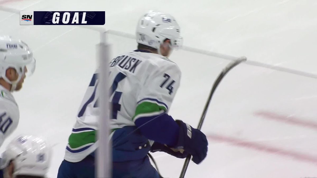Vancouver Canucks player Jake DeBrusk, wearing #74, celebrates near the goal after scoring, with an SN "GOAL" graphic displayed on the broadcast. His teammates are visible nearby as the team reacts to his first goal.