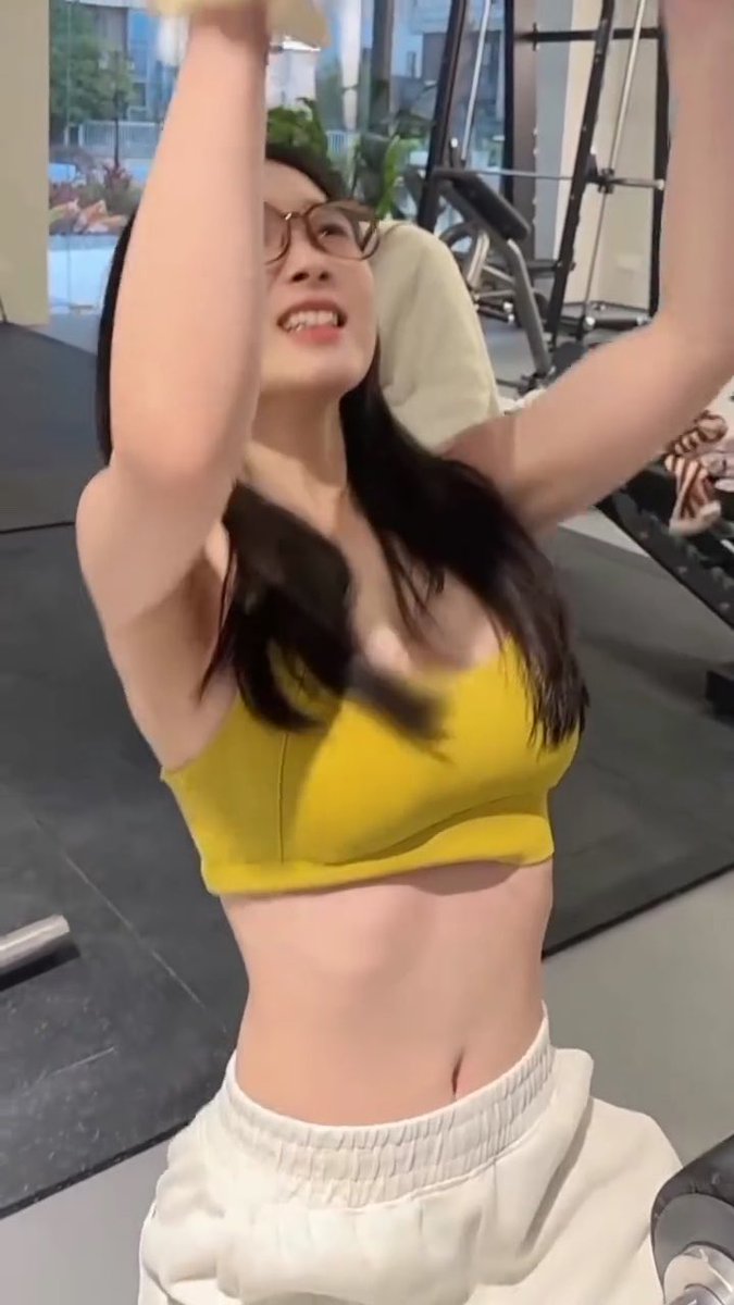 A woman in a yellow sports bra and white sweatpants works out at the gym, smiling with her arms raised overhead while using a piece of exercise equipment, exemplifying the post's caption about the motivating "eye candy" found in fitness spaces.