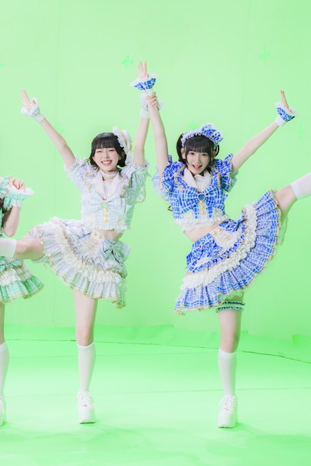 Two performers in elaborate blue and green frilled idol costumes pose energetically with one leg raised and arms linked on a bright green screen set, matching the cheerful "Ibuki Nemu Gymnastics" theme with their playful, dance-like stance.