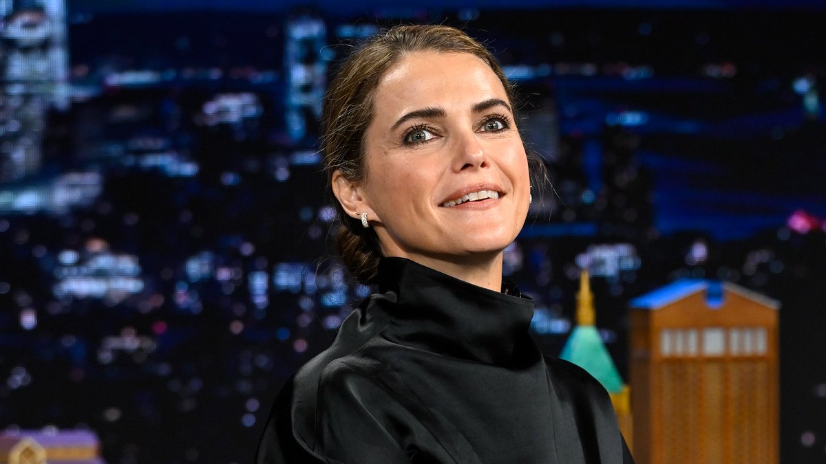 Emma Watson smiles warmly while wearing a high-necked black blouse and diamond earrings on a late-night talk show set, with a city skyline backdrop. Her genuine, joyful expression in the interview setting matches the post's tone of admiration for her candid, authentic personality.