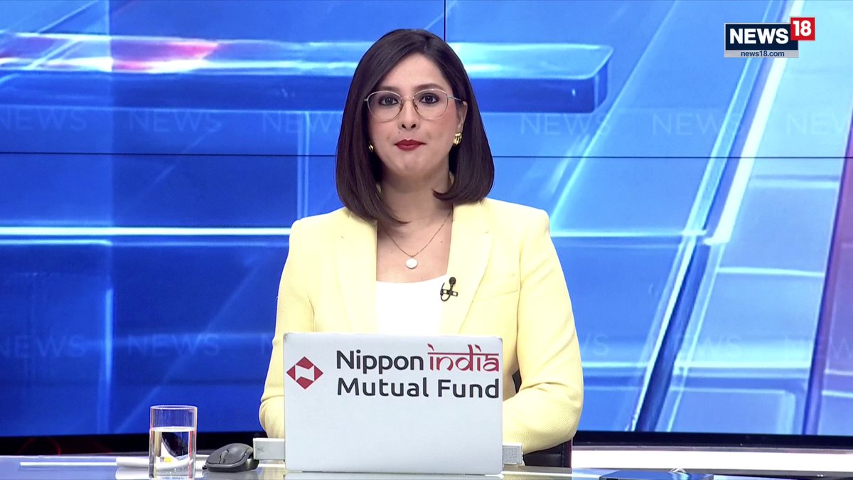 A female news anchor for News18, wearing a yellow blazer and glasses, sits at a desk with a Nippon India Mutual Fund-branded laptop and a glass of water. The accompanying post about US Green Card crackdowns and restrictions on Indian tech firms relates to a news broadcast she is likely anchoring on these topics.