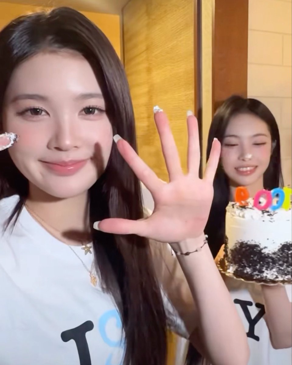 A selfie-style video of two young women celebrating with a cake decorated with colorful candles, with one woman waving at the camera.