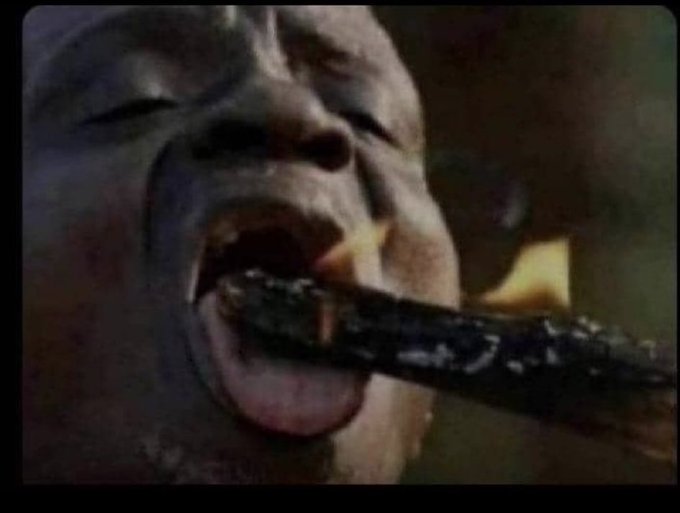 A close-up image from a video showing a person's open mouth with their tongue out, with a charred stick being inserted into their mouth.