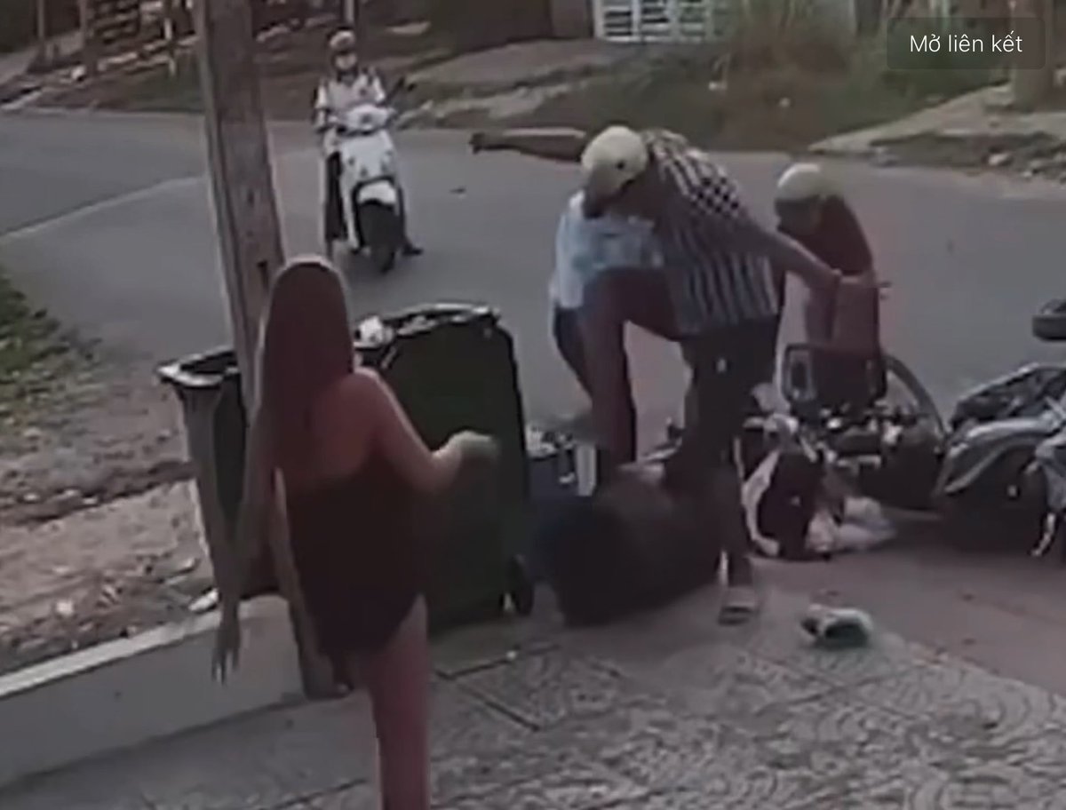 A CCTV street surveillance video still showing a chaotic scene involving multiple people and a motorbike on a street corner, with a woman in red walking away and the text 'Mở liên kết' visible in the top right.