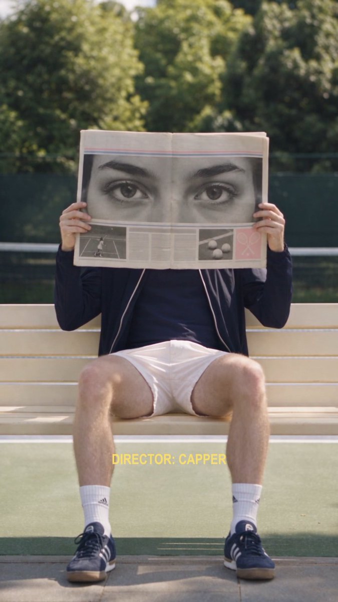 A person in shorts and sneakers sits on a wooden bench holding a newspaper up to cover their face, showing a close-up of a pair of eyes printed on the page, with 'DIRECTOR: CAPPER' text overlaid in yellow.