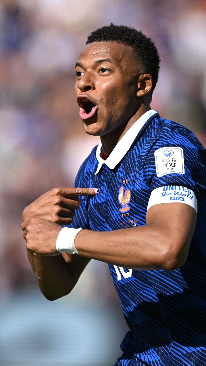 French footballer Kylian Mbappé, wearing the blue France national team jersey with a 'Unite for Peace FIFA' armband, points excitedly during a match.