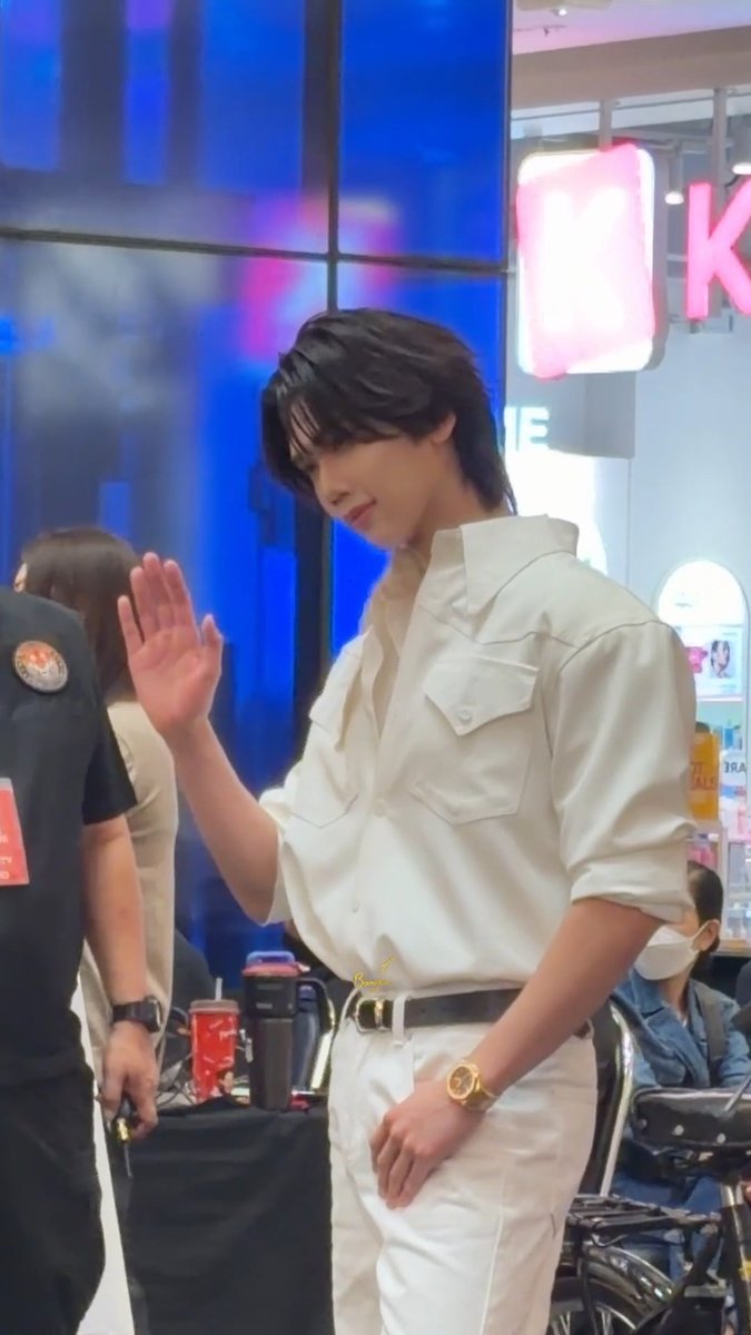 A young man with dark hair wearing a white shirt and white pants is waving his hand, appearing to be at an event with a security guard and staff nearby.