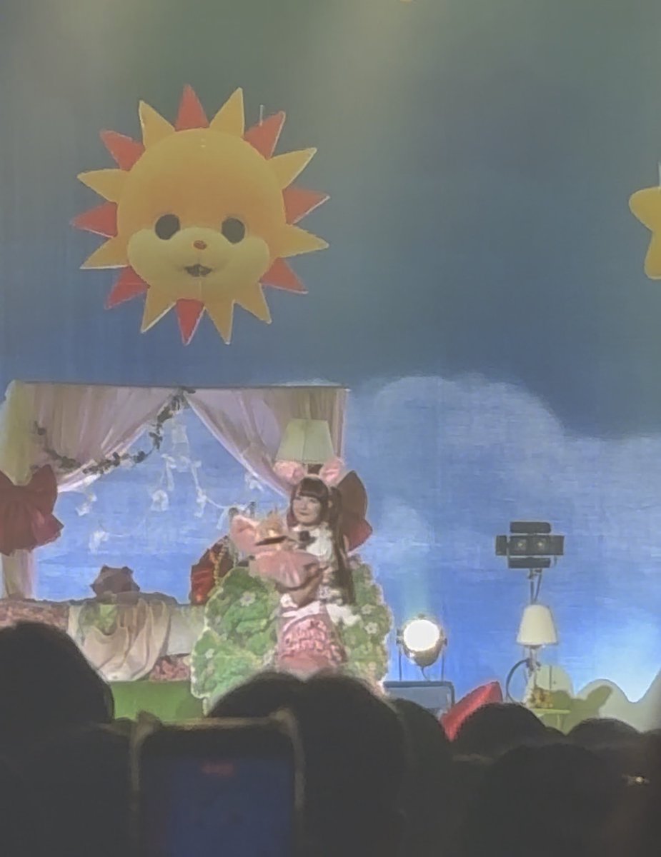 A person in elaborate costume performing on a stage decorated with a large smiling sun face, fairy lights, and lamps, viewed from the audience.