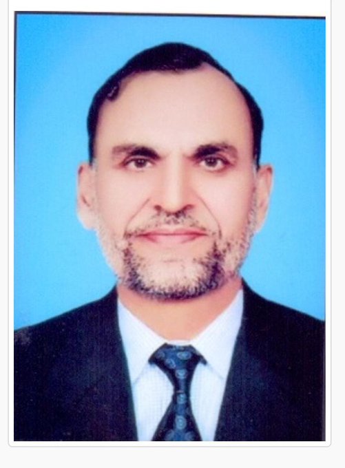 A formal portrait photograph of a man with short dark hair and a graying beard, wearing a dark suit, white shirt, and patterned tie against a blue background.