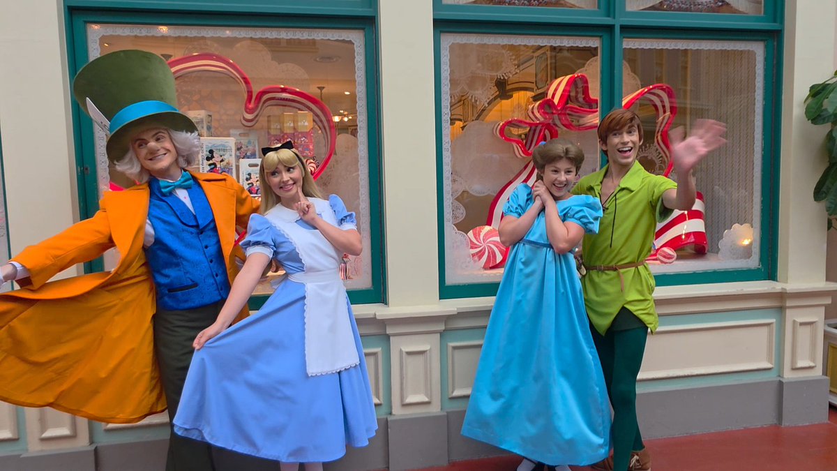 Four people in costumes portraying Alice in Wonderland characters—the Mad Hatter, Alice, another Alice, and Peter Pan—pose cheerfully in front of a decorative window display featuring candy-themed artwork.