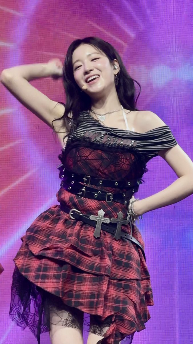 A smiling young woman performs on stage in a red and black plaid dress featuring leather belts with buckles, a striped off-shoulder neckline, and silver cross pendants, set against a purple background.
