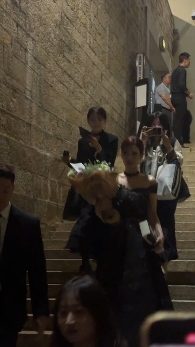 A woman in an elegant black off-shoulder dress holding a bouquet while descending a stone staircase, accompanied by others, including someone with a phone capturing the moment.