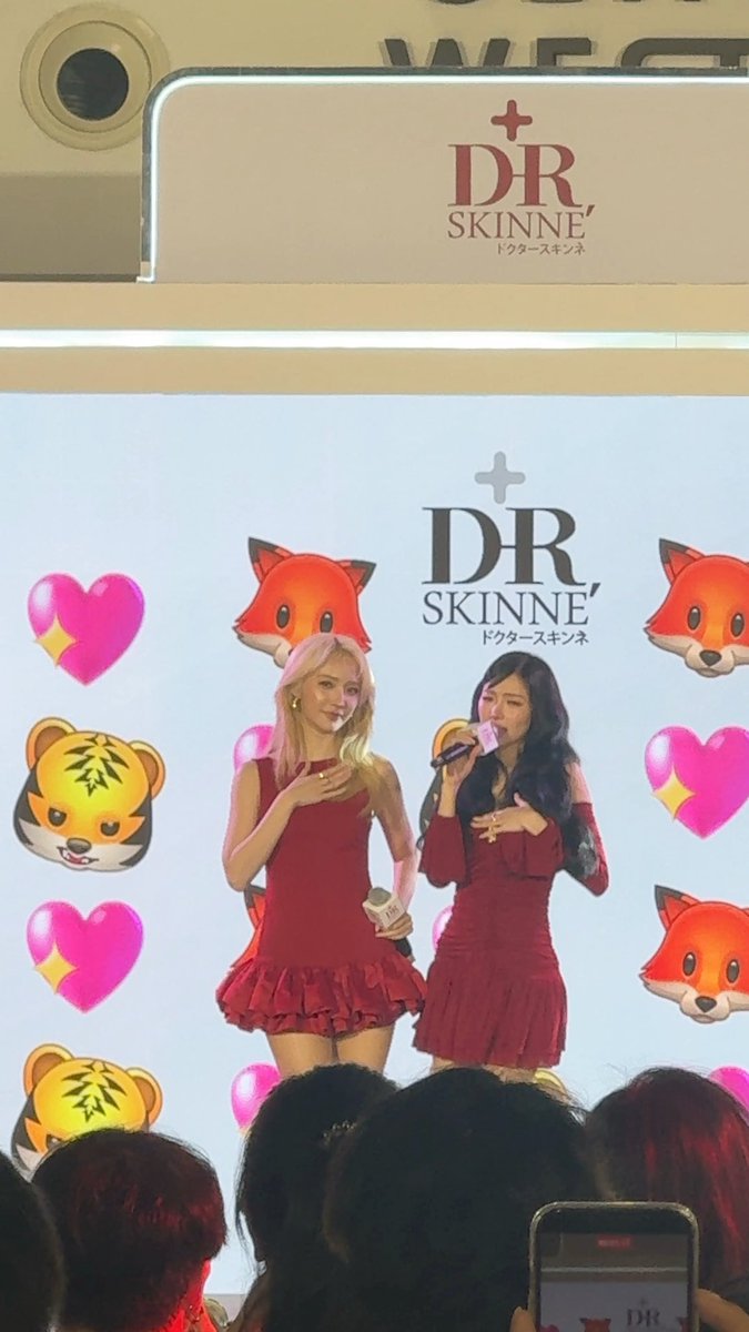 Two women in matching red ruffled dresses speak on stage at a DR SKINNE' (ドクタースキン) promotional event with a backdrop featuring fox and tiger animal emojis and pink hearts.