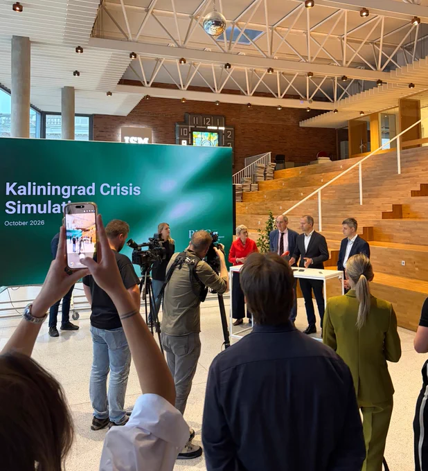 Press conference in the atrium of ISM University of Management and Economics, Vilnius. Journalists and camera operators film a panel standing at a white table, beside a large green screen reading “Kaliningrad Crisis Simulation, October 2026”. In the foreground a woman holds up a phone to film.
