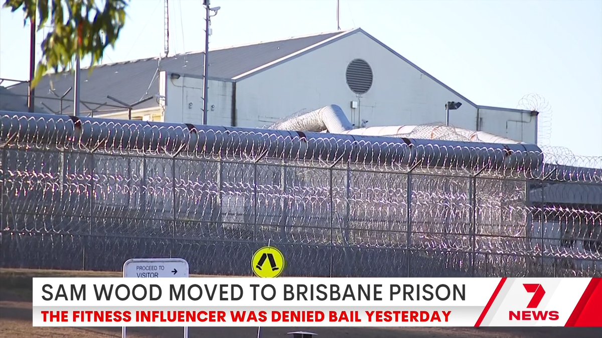A view of the Brisbane prison surrounded by razor wire, with a sign reading "PROCEED TO VISITOR." The image accompanies a 7 News chyron announcing "SAM WOOD MOVED TO BRISBANE PRISON - THE FITNESS INFLUENCER WAS DENIED BAIL YESTERDAY."